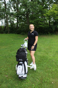 A woman in a black polo shirt and black skirt stands on a grassy golf course next to a white and black golf bag, with a black backpack attached. 